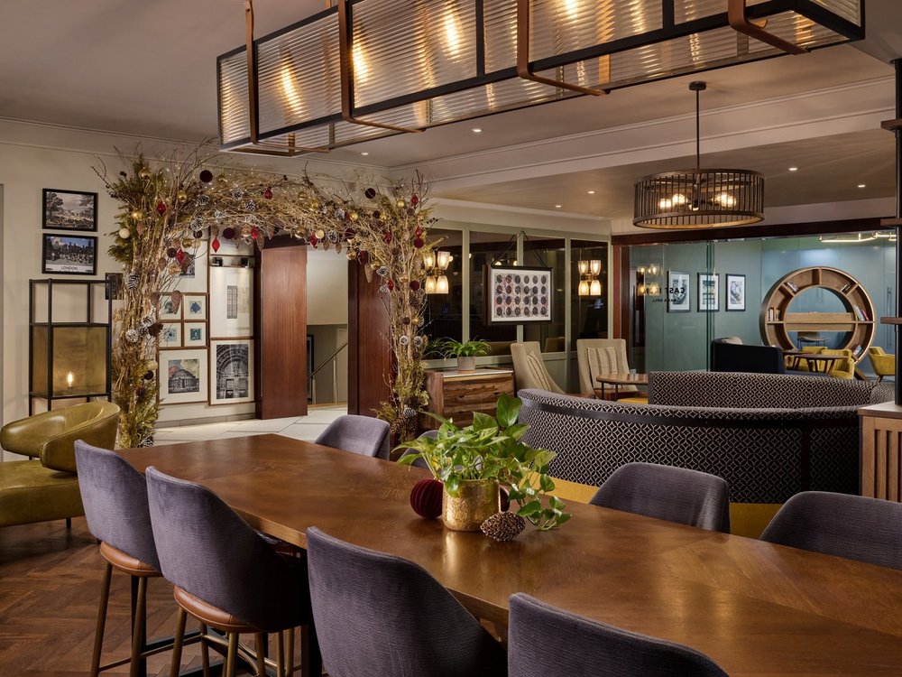 Interior view of the Cast Iron Bar & Grill at the London Marriott Hotel Kensington, featuring a long wooden dining table with blue upholstered chairs, modern hanging light fixtures, and a warm, inviting lounge seating area in the background.