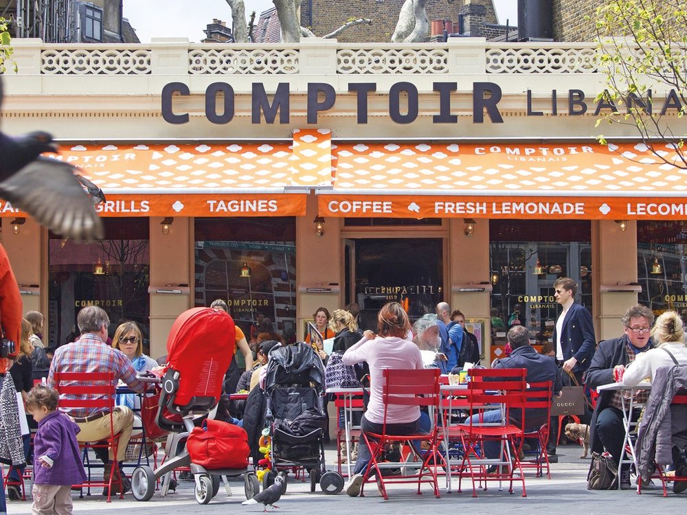 Patrons: Numerous people sit at small tables, talking and dining.Furniture: The patio is packed with vibrant red metal chairs and small round tables.Strollers: A bright red stroller and a black stroller are parked amidst the diners.