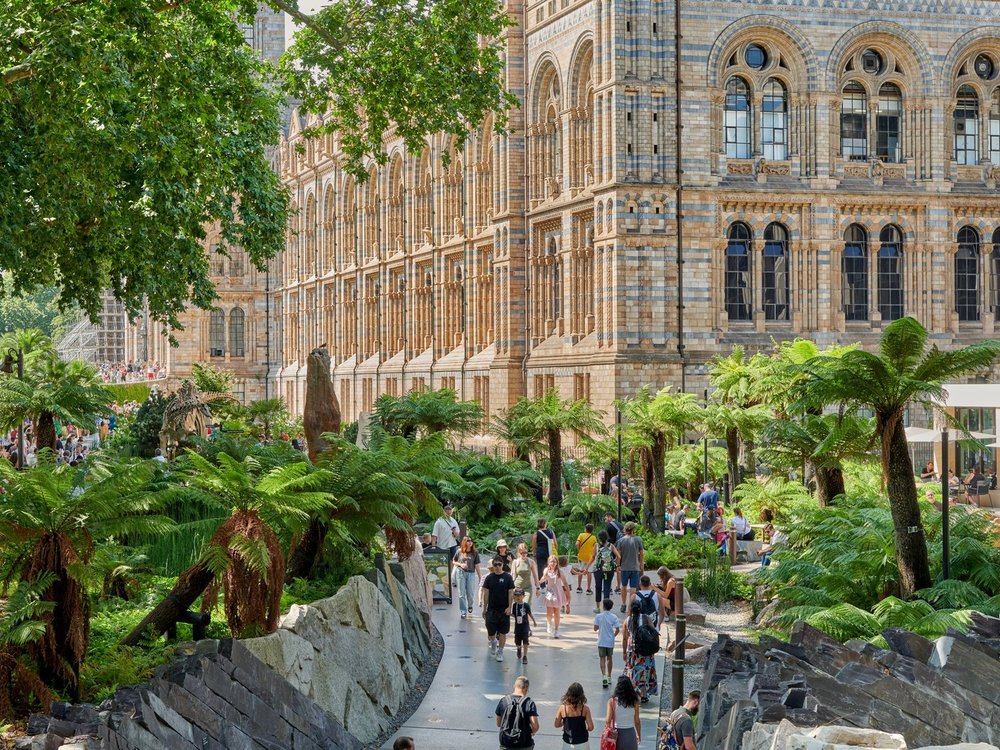A busy garden walkway beside a large, ornate museum building. Visitors stroll among lush ferns and rocks, with a dinosaur skeleton display visible and a café seating area nearby on a sunny day.