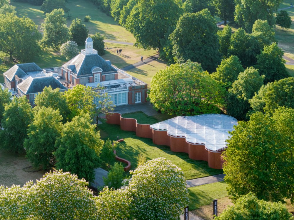 Aerial view of a historic brick building in a tree-filled park, beside a modern wavy-roof pavilion on a green lawn, with paths and a few people walking nearby.