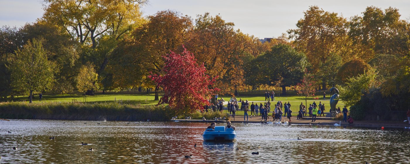A blue pedal boat on the Serpentine lake in Hyde Park during autumn, with people walking along the shore and vibrant fall foliage on the trees in the background.