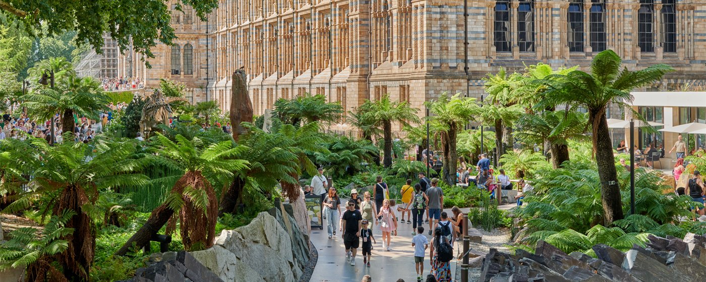 A busy garden walkway beside a large, ornate museum building. Visitors stroll among lush ferns and rocks, with a dinosaur skeleton display visible and a café seating area nearby on a sunny day.