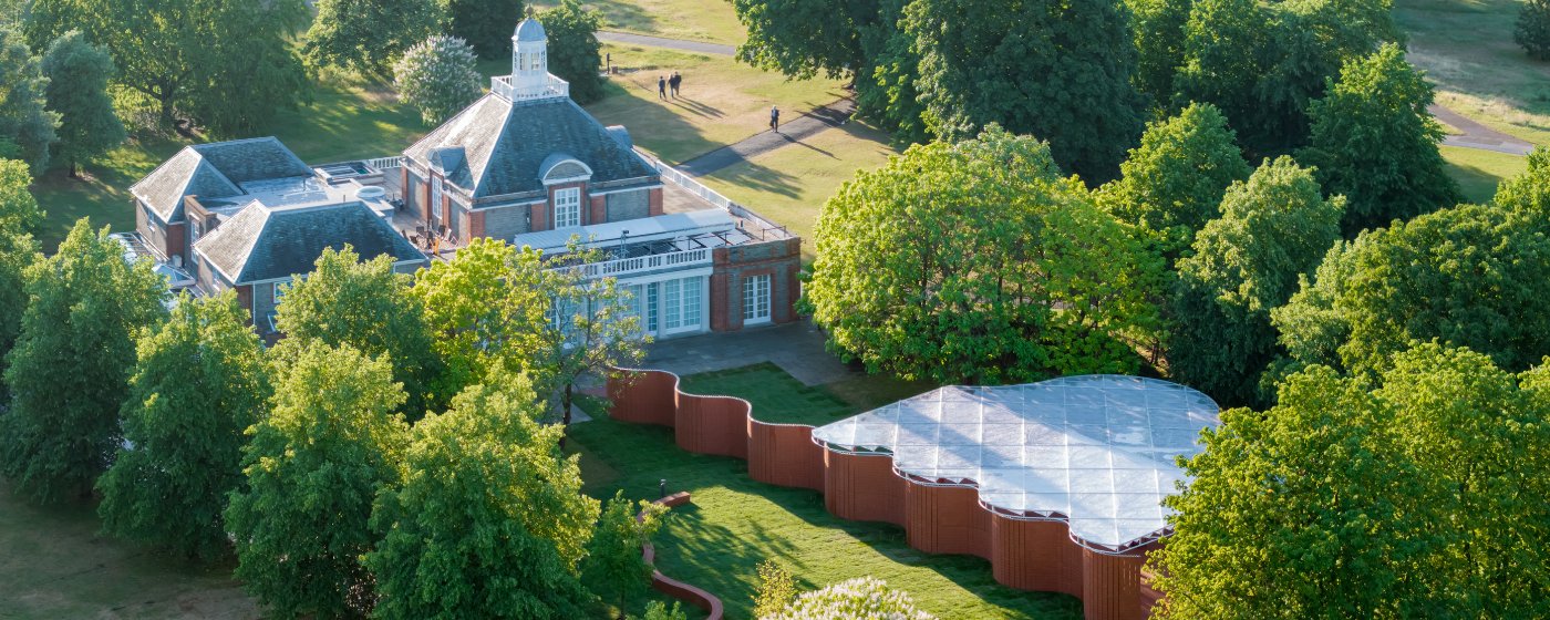 Aerial view of a historic brick building in a tree-filled park, beside a modern wavy-roof pavilion on a green lawn, with paths and a few people walking nearby.