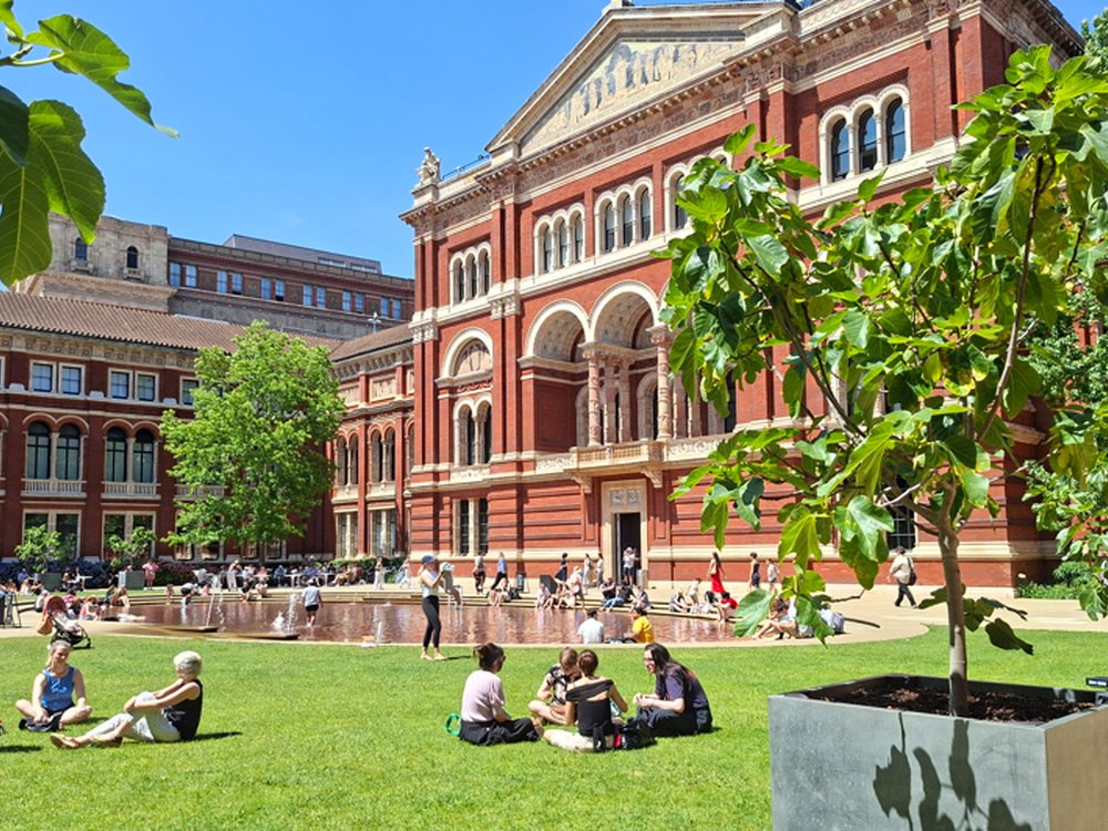 Visitors relaxing on the lawn and around the shallow fountain in the John Madejski Garden, with the red-brick façade of the V&A South Kensington behind them on a sunny day.