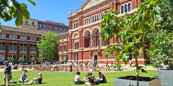 Visitors relaxing on the lawn and around the shallow fountain in the John Madejski Garden, with the red-brick façade of the V&A South Kensington behind them on a sunny day.