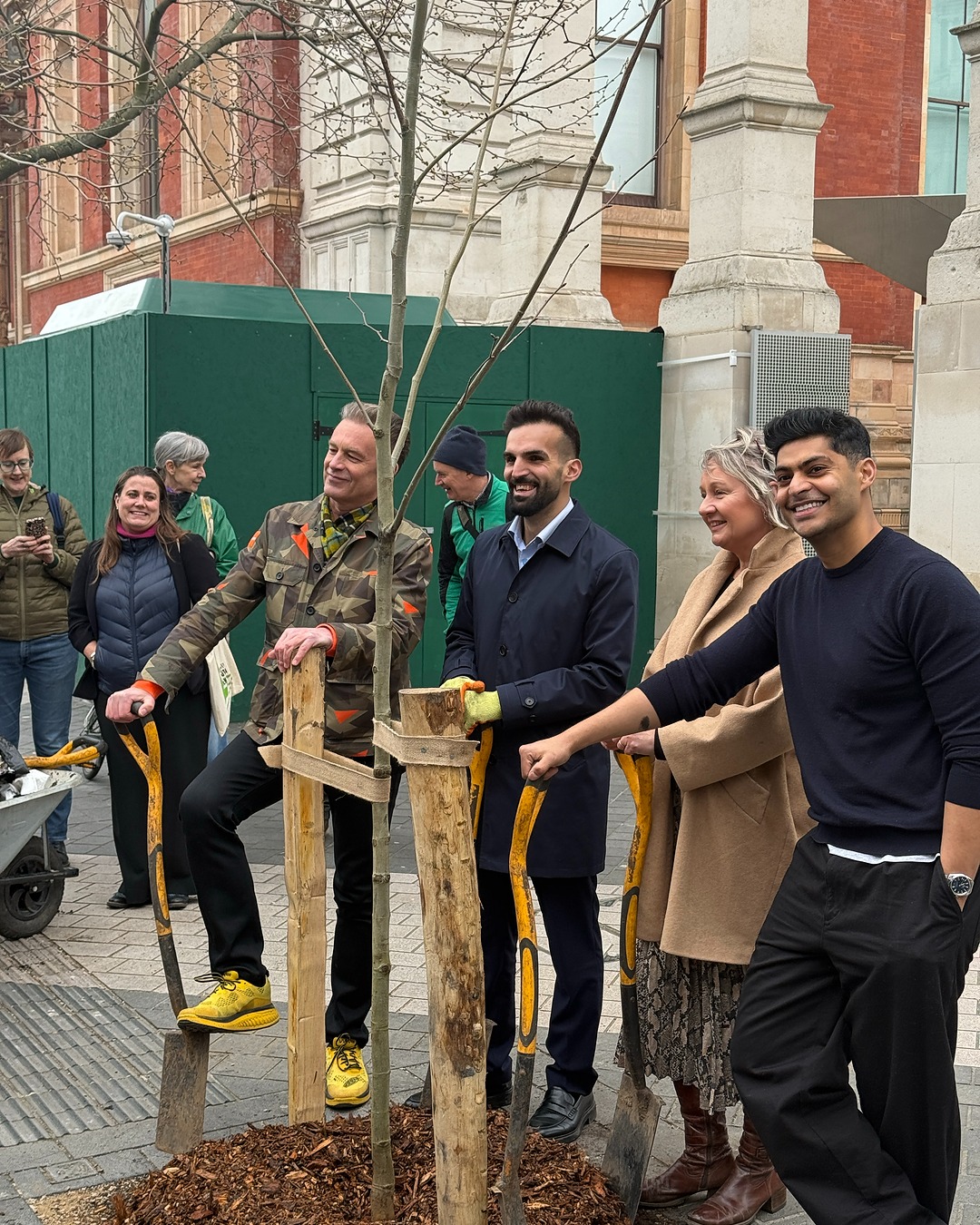 Planted plane tree on Exhibition Road Participants gathered around a newly planted tree in an urban square, posing beside the young sapling supported by wooden stakes.