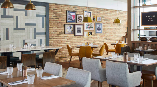 Modern restaurant dining area with wooden tables, upholstered chairs, pendant lighting and framed artwork displayed on an exposed brick wall.