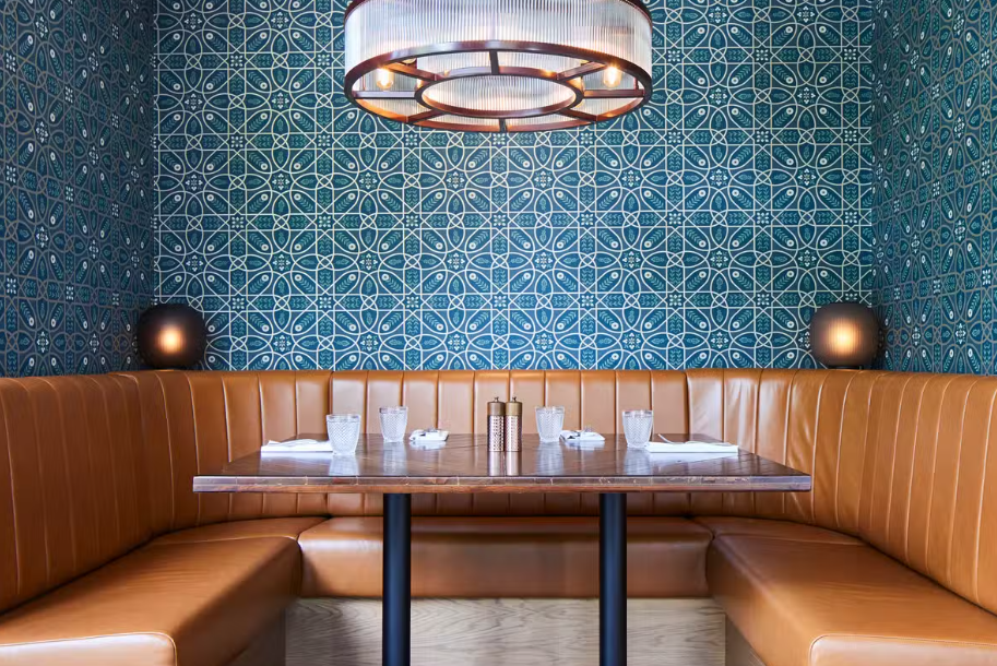 Restaurant booth seating with tan leather banquettes, a wooden dining table and a blue geometric-patterned feature wall under a pendant light.