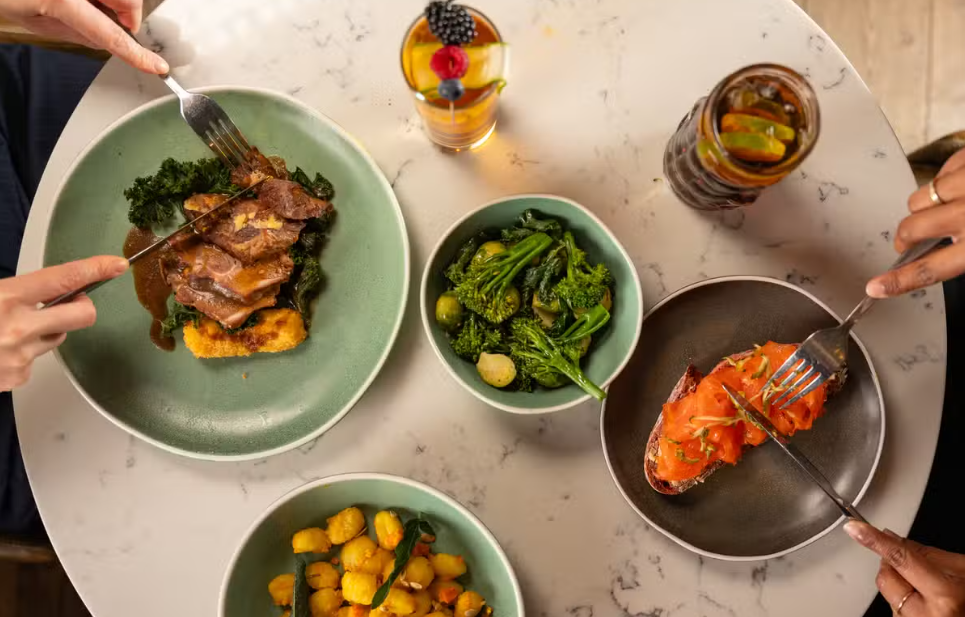 Overhead view of a restaurant table with plated dishes including grilled meat, lobster, vegetables and drinks, with diners serving themselves