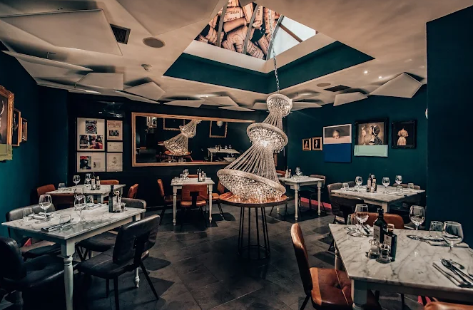 Restaurant dining room with wooden tables, upholstered chairs and framed artwork on an exposed brick wall.