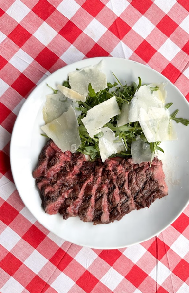 Plate of sliced grilled steak served with rocket leaves and shaved Parmesan cheese on a red-and-white checkered tablecloth.