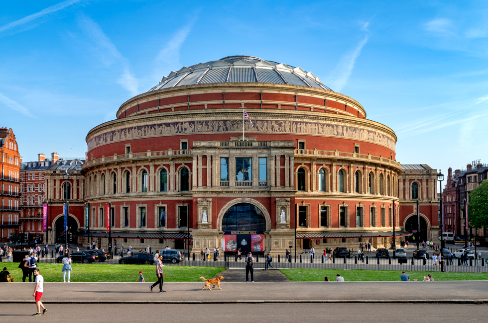 Exterior view of the Royal Albert Hall with its iconic circular red-brick facade and domed roof, seen from the park with people walking in the foreground.