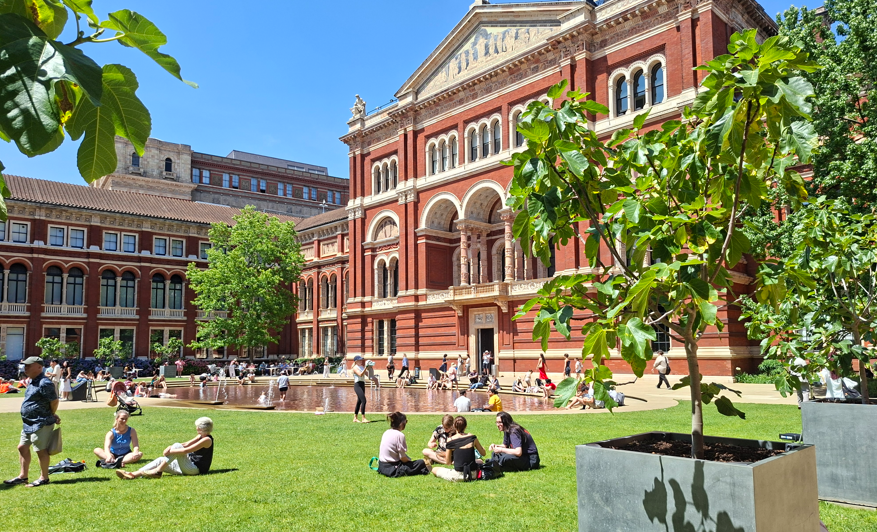 John Madejski Garden Visitors relaxing on the lawn and around the shallow fountain in the John Madejski Garden, with the red-brick façade of the V&A South Kensington behind them on a sunny day.