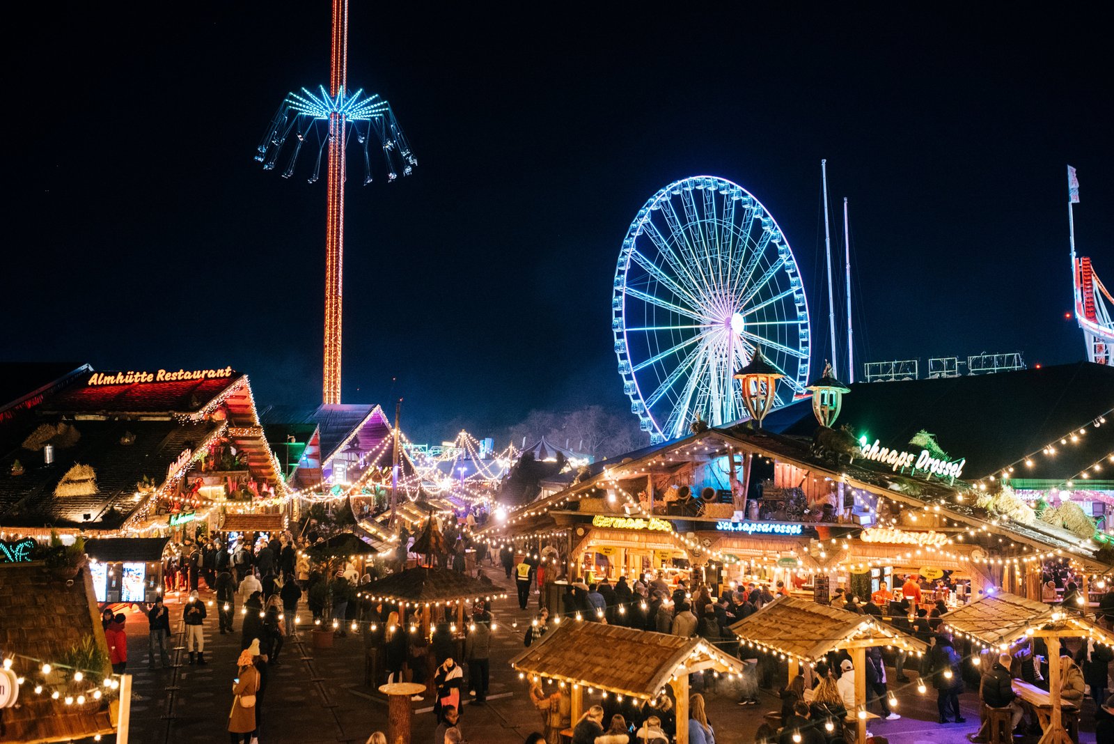 A nighttime view of Hyde Park Winter Wonderland featuring illuminated market-style chalets, festive crowds, thrill rides, and a large Ferris wheel glowing against the night sky.
