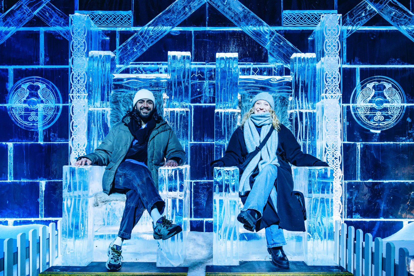 Two visitors sit on intricately carved ice thrones inside the Magical Ice Kingdom at Hyde Park Winter Wonderland, surrounded by glowing blue ice sculptures and frozen architectural details.