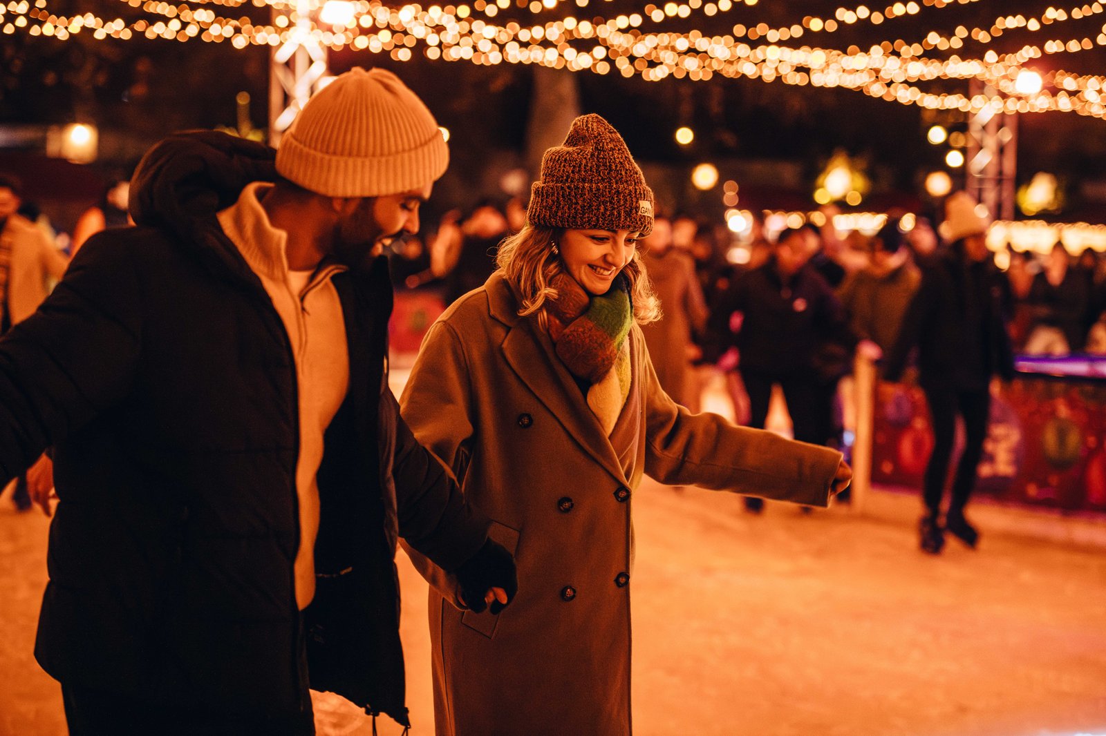 Two visitors skate hand in hand across the outdoor ice rink at Hyde Park Winter Wonderland, with festive lights twinkling overhead and other skaters in the background.