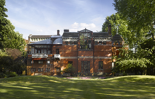 Historic red-brick museum building viewed across a large green lawn, with trees framing the scene and glass structures visible on the roof.